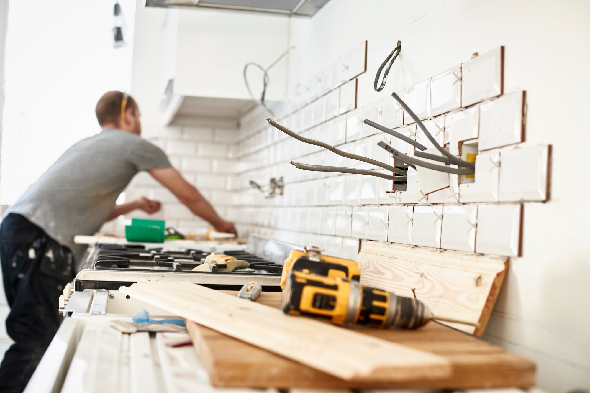 A man working in a new kitchen, a tiler applying tiles to the wall behind the cooker.